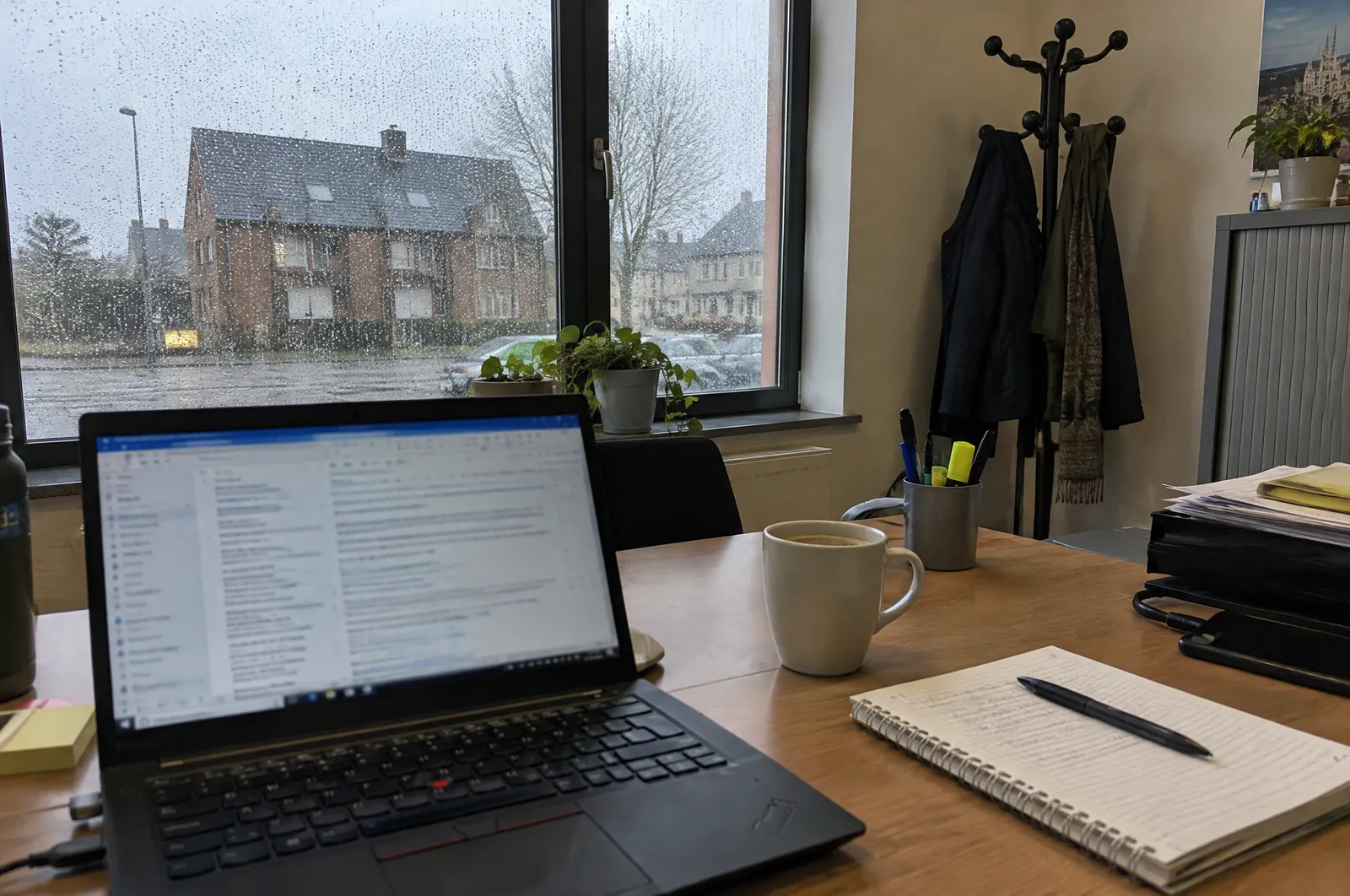 Een laptop met een mailprogramma op een bureau in een klein kantoor, naast een notitieblok en een kop koffie