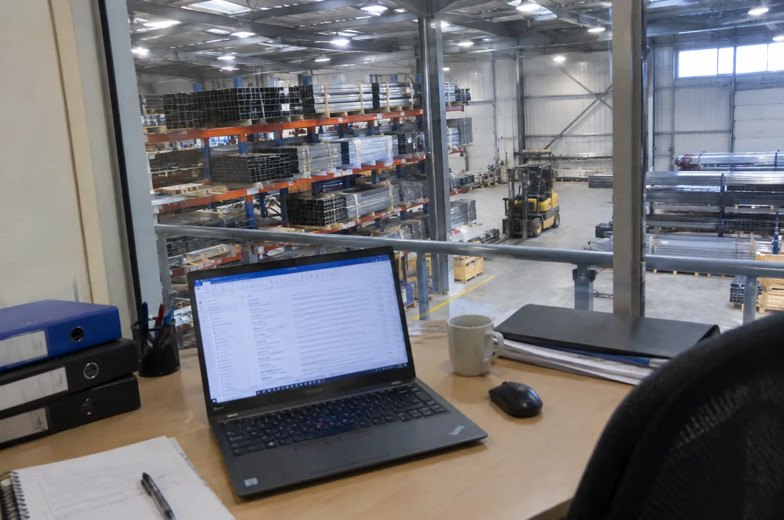 Een kantoorbureau met een laptop met een volle inbox, een stapel ordners en een kop koffie, met door het raam een werkplaats