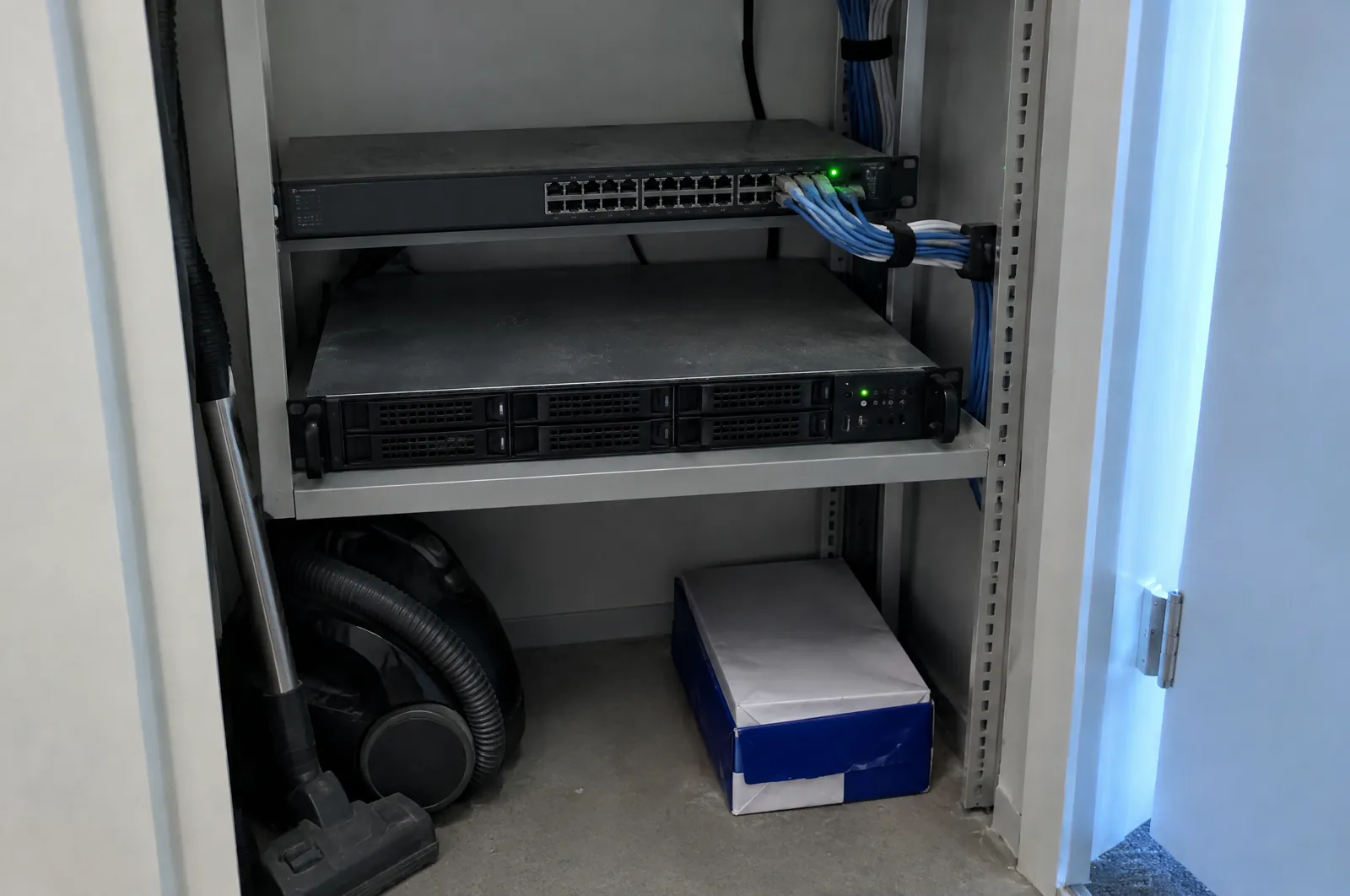 Kleine server in een rek in een opgeruimd technisch kastje, met netwerkkabels en een groen statuslampje