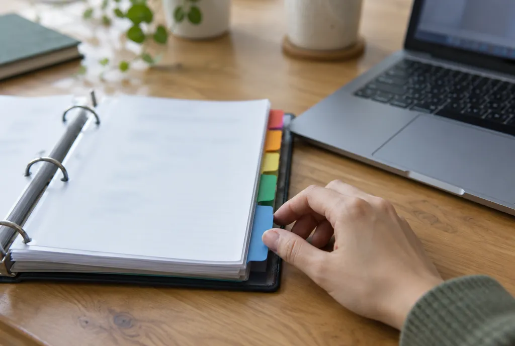 Een open ordner met tabbladen naast een laptop op een houten bureau